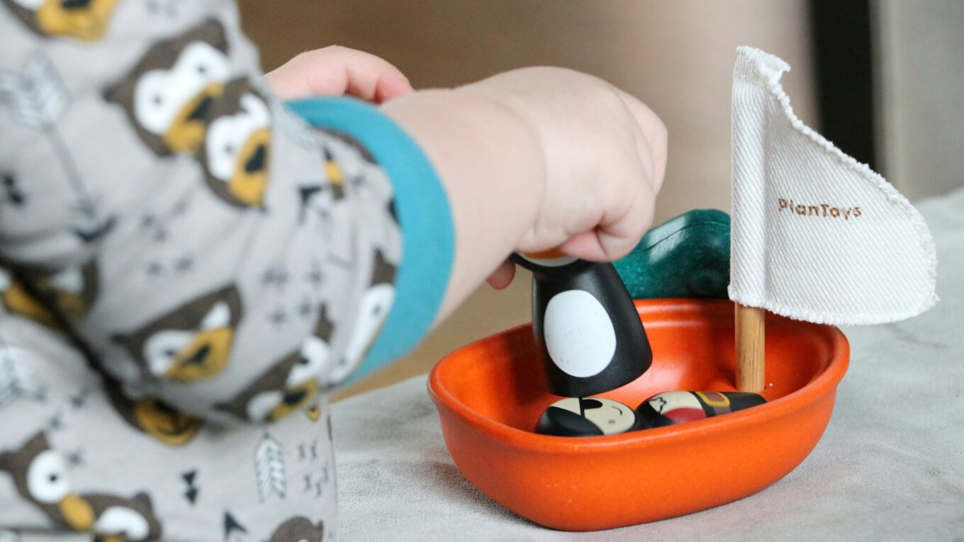 "Close-up of a child wearing owl-patterned pajamas, playing with a small orange toy boat featuring wooden figures and a fabric sail labeled 'PlanToys'."