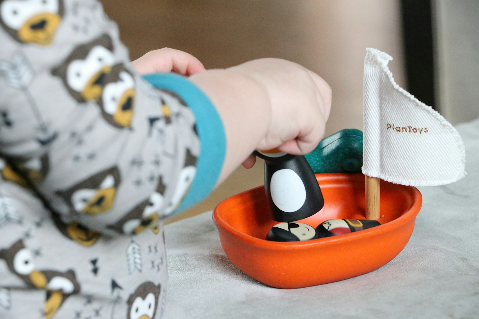 "Close-up of a child wearing owl-patterned pajamas, playing with a small orange toy boat featuring wooden figures and a fabric sail labeled 'PlanToys'."